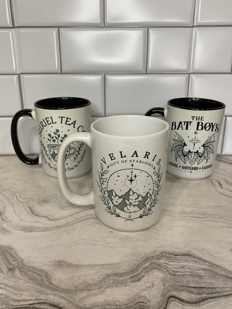 Puede incluir: Tres tazas de cer&aacute;mica blanca con asas negras y borde negro. Las tazas tienen dise&ntilde;os diferentes. La primera taza tiene una ilustraci&oacute;n en blanco y negro de una taza de t&eacute; y flores con el texto "RIEL TEA C" y "Tea In Prythian". La segunda taza tiene una ilustraci&oacute;n en blanco y negro de una cordillera con estrellas y el texto "VELARIS" y "CITY OF STARLIGHT". La tercera taza tiene una ilustraci&oacute;n en blanco y negro de un murci&eacute;lago con el texto "THE BAT BOYS" y "AIRIEL + RHYSAND + CASCAR".
