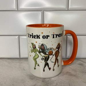 May include: White ceramic mug with an orange rim and handle. The mug features a black and white illustration of three women dressed in Halloween costumes. The text "Trick or Treat" is printed in black on the mug.