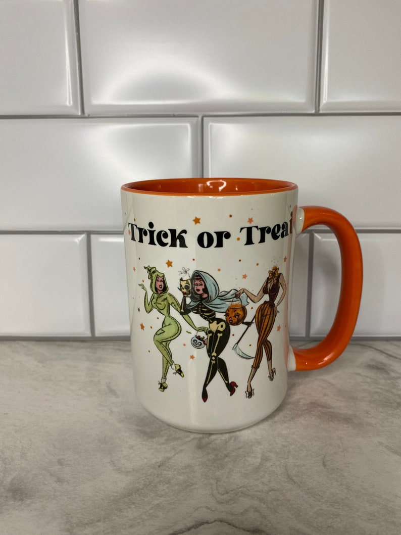 May include: White ceramic mug with an orange rim and handle. The mug features a black and white illustration of three women dressed in Halloween costumes. The text "Trick or Treat" is printed in black on the mug.