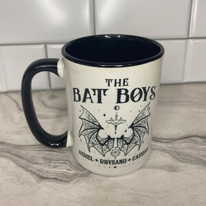 Puede incluir: Taza de cer&aacute;mica blanca con asa y borde negros. La taza tiene un gr&aacute;fico negro de alas de murci&eacute;lago con el texto "THE BAT BOYS" y los nombres "AZRIEL, RHYSAND, CASSIAN" debajo.