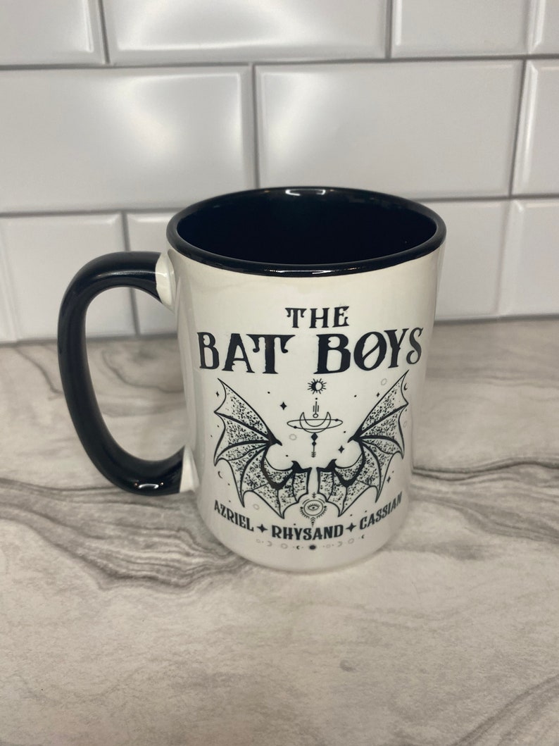 Puede incluir: Taza de cer&aacute;mica blanca con asa y borde negros. La taza tiene un gr&aacute;fico negro de alas de murci&eacute;lago con el texto "THE BAT BOYS" y los nombres "AZRIEL, RHYSAND, CASSIAN" debajo.