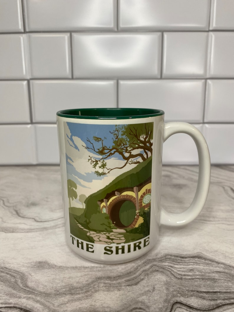 May include: White ceramic mug with a green rim. The mug features a colorful illustration of a hobbit hole with a green door and a stone path leading to it. The text "THE SHIRE" is printed on the bottom of the mug.