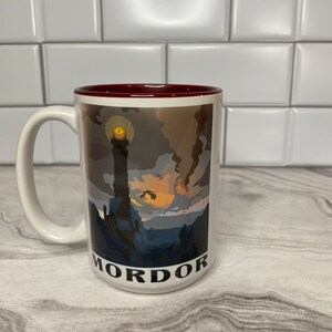 May include: White ceramic mug with a red rim. The mug features a graphic of a dark, mountainous landscape with a tall tower and a glowing eye. The text "Mordor" is printed on the bottom of the mug.