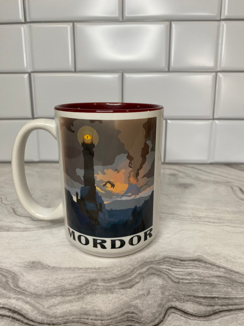 May include: White ceramic mug with a red rim. The mug features a graphic of a dark, mountainous landscape with a tall tower and a glowing eye. The text "Mordor" is printed on the bottom of the mug.