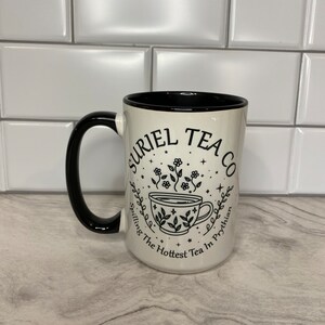 Puede incluir: Taza de cer&aacute;mica blanca con asa negra y texto negro que dice "Uriel Tea Co.  Spilling The Hottest Tea In Prythian" con una ilustraci&oacute;n estilizada de una taza de t&eacute; con vapor y flores.
