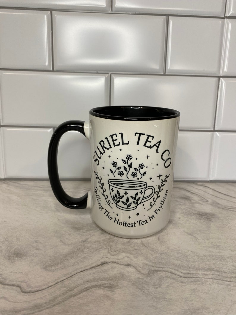 Puede incluir: Taza de cer&aacute;mica blanca con asa negra y texto negro que dice "Uriel Tea Co.  Spilling The Hottest Tea In Prythian" con una ilustraci&oacute;n estilizada de una taza de t&eacute; con vapor y flores.