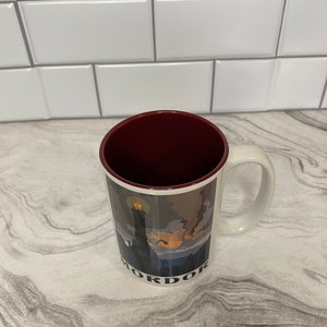 May include: White ceramic mug with a red rim. The mug has a graphic of a dark landscape with a tower and the word "Mordor" printed on the bottom.