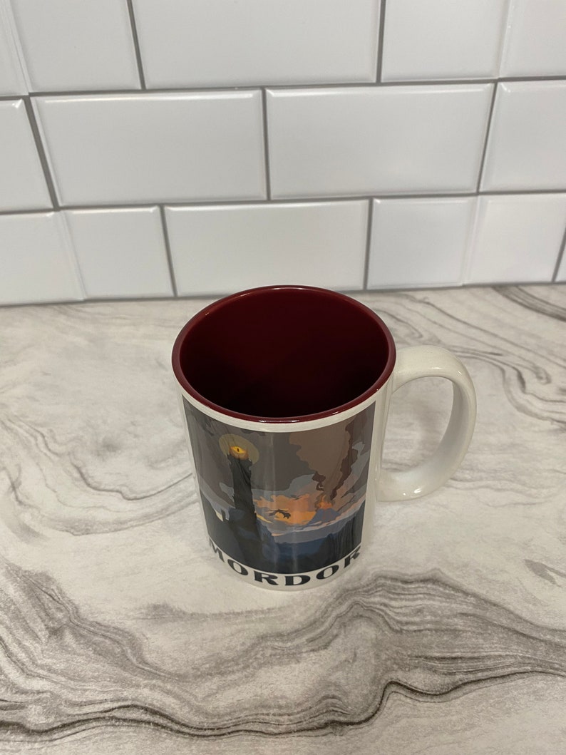 May include: White ceramic mug with a red rim. The mug has a graphic of a dark landscape with a tower and the word "Mordor" printed on the bottom.