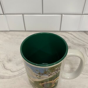 May include: A white ceramic mug with a green interior and a handle. The mug features a colorful illustration of a hobbit hole with the text "The Shire" at the bottom.