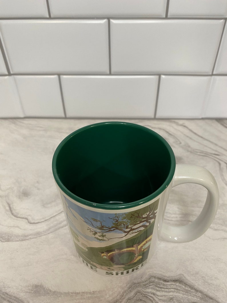 May include: A white ceramic mug with a green interior and a handle. The mug features a colorful illustration of a hobbit hole with the text "The Shire" at the bottom.