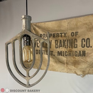 May include: A vintage metal light fixture shaped like a dough hook, with a light bulb inside. The fixture is hanging from a black cord. A burlap banner behind the fixture reads "Property of Wesley Baking Co. Detroit, Michigan".