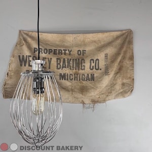 Può includere: Una lampada a sospensione realizzata con una frusta in metallo argentato, dotata di una lampadina centrale in stile Edison. La lampada pende da un cavo nero davanti a un sacco di tessuto color cuoio con la scritta PROPERTY OF WESLEY BAKING CO. MICHIGAN.