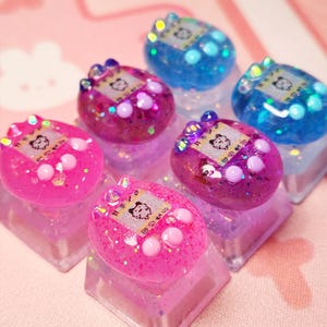 Puede incluir: Primer plano de seis teclas de teclado coloridas y brillantes. Cada tecla tiene la forma de un juego portátil retro, con una pequeña pantalla y botones. Las teclas son en tonos de rosa y azul, con una base transparente y brillante. El fondo es una tela rosa claro.