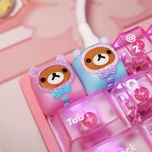 May include: Close-up of two bear-shaped keycaps in pink and blue, with brown faces and black eyes. The keycaps are on a pink keyboard, with transparent pink keys visible. The keycaps have a bow tie and button details.