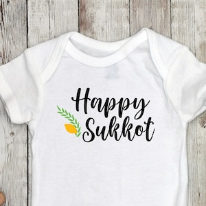 Puede incluir: Un body blanco para bebé con el texto "Happy Sukkot" en letras negras y un gráfico de citron verde y amarillo.