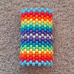 May include: A rainbow-colored beaded bracelet with a white and purple center. The bracelet is made of small plastic beads and is about 2 inches wide.
