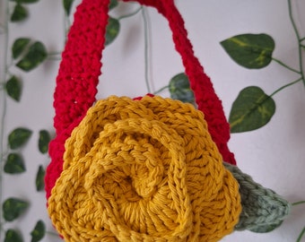 May include: A crocheted headband with a large yellow rose and green leaves. The headband is red.