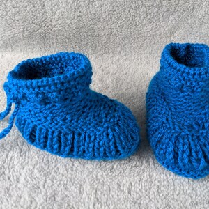 May include: A pair of handmade, bright blue crochet baby booties. The booties have a ribbed design around the soles and cuffs, with a drawstring detail on one. The soft, knitted texture suggests warmth and comfort, perfect for a newborn.