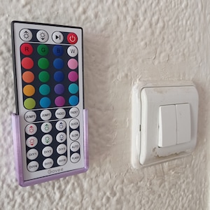 Peut inclure: Un mur blanc avec un support de télécommande violet fixé dessus. Le support contient une télécommande avec une variété de boutons, y compris des options de couleur, saut, fondu, rapide, lent, automatique, flash et paramètres DIY. La télécommande porte également l'inscription "Govee". Un interrupteur lumineux blanc se trouve à droite du support.