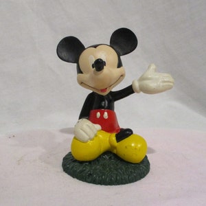 Puede incluir: Una figura de Mickey Mouse en blanco y negro sentada sobre una base verde. Mickey lleva pantalones cortos rojos y zapatos amarillos. Est&aacute; saludando con la mano derecha.