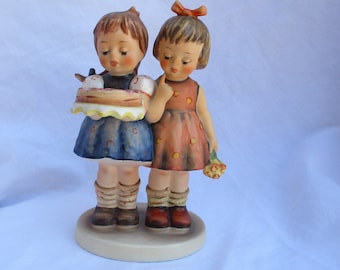 Puede incluir: Dos figuritas de porcelana de niñas, una sosteniendo un pastel y la otra una flor. La niña que sostiene el pastel lleva un vestido azul con lunares blancos y la niña que sostiene la flor lleva un vestido rojo con lunares amarillos.