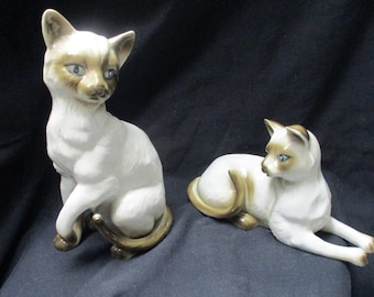May include: Two porcelain Siamese cat figurines. One cat is sitting with its tail curled around its paws. The other cat is lying down with its head turned to the side. Both cats have blue eyes and brown markings.