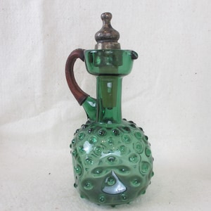 May include: A green glass bottle with a textured surface and a brown wooden handle. The bottle has a silver stopper and a round, bulbous shape.