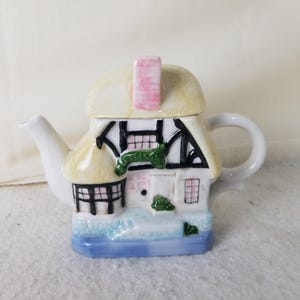 May include: A ceramic teapot shaped like a quaint cottage. The teapot features a thatched roof, black and white timber framing, and a pink chimney. The base is painted with a blue water scene, and the handle and spout are white.