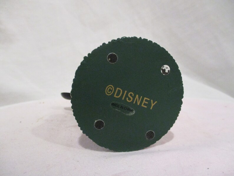 Puede incluir: La parte inferior de una figurita verde con un copyright de Disney en dorado y una etiqueta que dice "Hecho en China".
