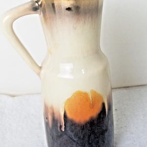 Puede incluir: Un jarrón de cerámica con asa, con un esmalte blanco, marrón y naranja. El jarrón tiene un borde dorado metálico.