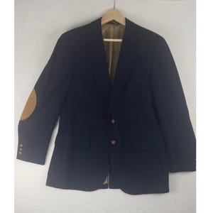 May include: A navy blue blazer with tan elbow patches and brown buttons. The blazer has two front pockets and is hanging on a wooden hanger. The interior lining is a light brown color.