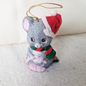 May include: A ceramic Christmas ornament shaped like a gray mouse wearing a red and white Santa hat and a green and red striped scarf. The mouse has pink ears and feet, and a gold string for hanging.