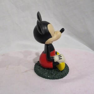 Puede incluir: Una figura de Mickey Mouse de color negro y rojo sentada sobre una base verde. Mickey lleva sus caracter&iacute;sticos pantalones cortos rojos y zapatos amarillos. Tiene la nariz negra y orejas grandes.