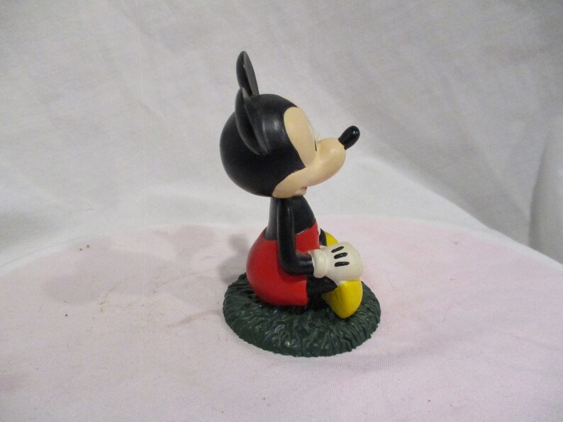 Puede incluir: Una figura de Mickey Mouse de color negro y rojo sentada sobre una base verde. Mickey lleva sus caracter&iacute;sticos pantalones cortos rojos y zapatos amarillos. Tiene la nariz negra y orejas grandes.