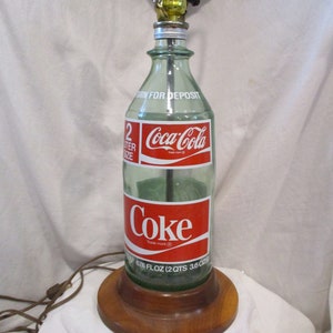 May include: A vintage Coca-Cola bottle lamp with a wooden base. The bottle is clear glass with a red and white label that says "Coca-Cola" and "Coke". The label also says "676 FLOZ (2QTS 36 OZ)".