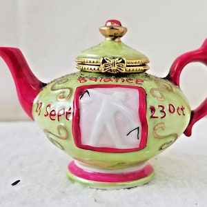 Può includere: Una piccola teiera verde e rossa con un coperchio dorato e un simbolo zodiacale bianco all'interno. La teiera ha le parole "23 Ott" e "27 Set" sul lato e la parola "Balance" sul coperchio.