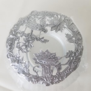 Pode incluir: Um prato decorativo prateado com um intrincado design floral e arquitetônico. O prato apresenta entalhes detalhados de flores, folhas e um pequeno edifício. O prato está envolto em plástico transparente.