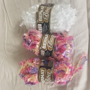 May include: Three skeins of Bernat Boa yarn. The top skein is solid white. The middle and bottom skeins feature a variegated blend of pink, purple, and yellow. Each skein has a black paper label with white text and a weight of 1.75 ounces.