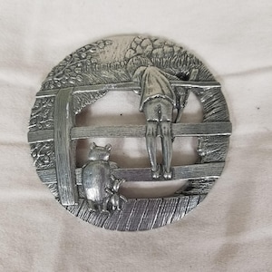 Könnte beinhalten: Eine kreisförmige, silberfarbene Metallplakette mit einer Reliefdarstellung einer Person, die sich über einen Holzzaun lehnt, während ein Bär und ein kleines schweineartiges Wesen daneben stehen. Das Design zeigt strukturiertes Laub und Holzdielen.
