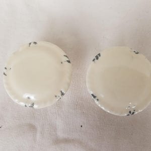 Puede incluir: Dos cuencos de cerámica blanquecina con forma de concha y motas gris oscuro en los bordes. Los cuencos son redondos y tienen un borde ligeramente elevado. Están colocados sobre una superficie clara.