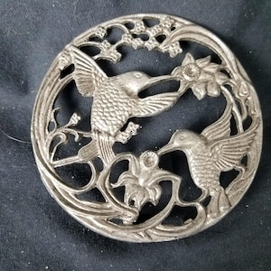 以下が含まれることがあります： 2 羽のハチドリと花のディテールが特徴の、円形の銀色の金属製ブローチ。鳥は翼を広げ、装飾的な透かし彫りのフレームの中で花と交流している様子が描かれています。ブローチはヴィンテージ感があります。