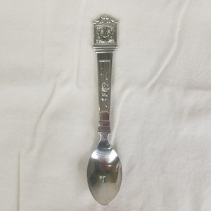 May include: A silver-colored spoon with an ornate handle. The handle features a decorative design with a clock-like shape at the top and engraved details along the stem. The spoon's bowl is oval-shaped and reflects the surrounding environment.