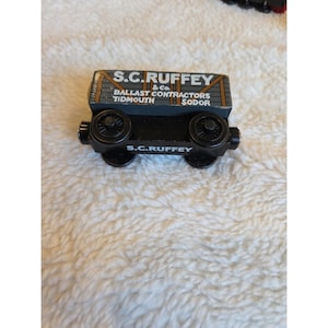 Puede incluir: Un vagón de tren de juguete de madera, negro con techo gris y detalles marrones, con el texto "S.C. Ruffey & Co. Ballast Contractors Tidmouth Sodor". Las ruedas son negras.