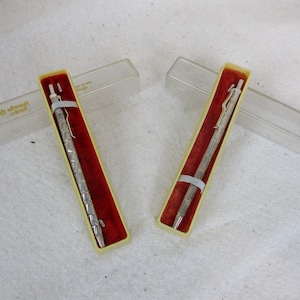 May include: Two silver pens in their original boxes. The pens have a textured design and are in clear plastic boxes with red velvet lining.