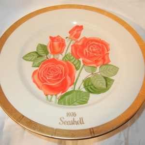 Può includere: Un piatto in ceramica bianca con un bordo dorato presenta un motivo floreale di tre rose arancioni con foglie verdi. Il piatto ha il testo "1976 Seashell" in lettere dorate.