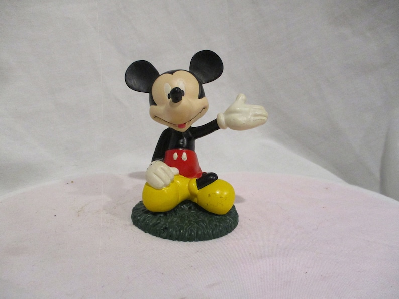 Puede incluir: Una figura de Mickey Mouse en blanco y negro sentada sobre una base verde. Mickey lleva pantalones cortos rojos y zapatos amarillos. Est&aacute; saludando con la mano derecha.