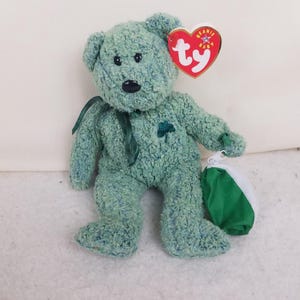 May include: A green plush bear with a shamrock on its chest and a green ribbon around its neck. The bear has a red heart-shaped tag with the word "TY" on it. A small green and white pouch is next to the bear.