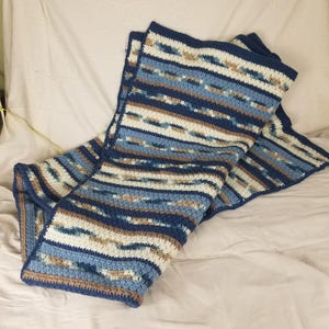 May include: A rectangular crochet blanket featuring horizontal stripes in shades of navy blue, light blue, cream, and tan. The pattern consists of alternating solid-colored rows and textured, variegated rows that create a repeating wave-like design.