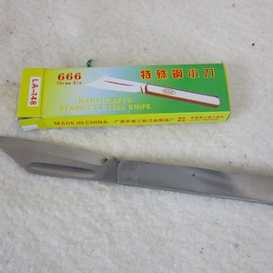 May include: A small, silver folding knife with a green and yellow box. The box has the text "666 Three Six HANDICRAFTS STAINLESS STEEL KNIFE MADE IN CHINA" and "特殊钢小刀" in Chinese characters. The box also has the text "LA-148".