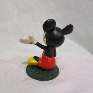 Puede incluir: Una peque&ntilde;a figura de Mickey Mouse de color negro y rojo sentada sobre una base verde. Mickey lleva sus caracter&iacute;sticos pantalones cortos rojos y zapatos amarillos. Est&aacute; saludando con la mano derecha.